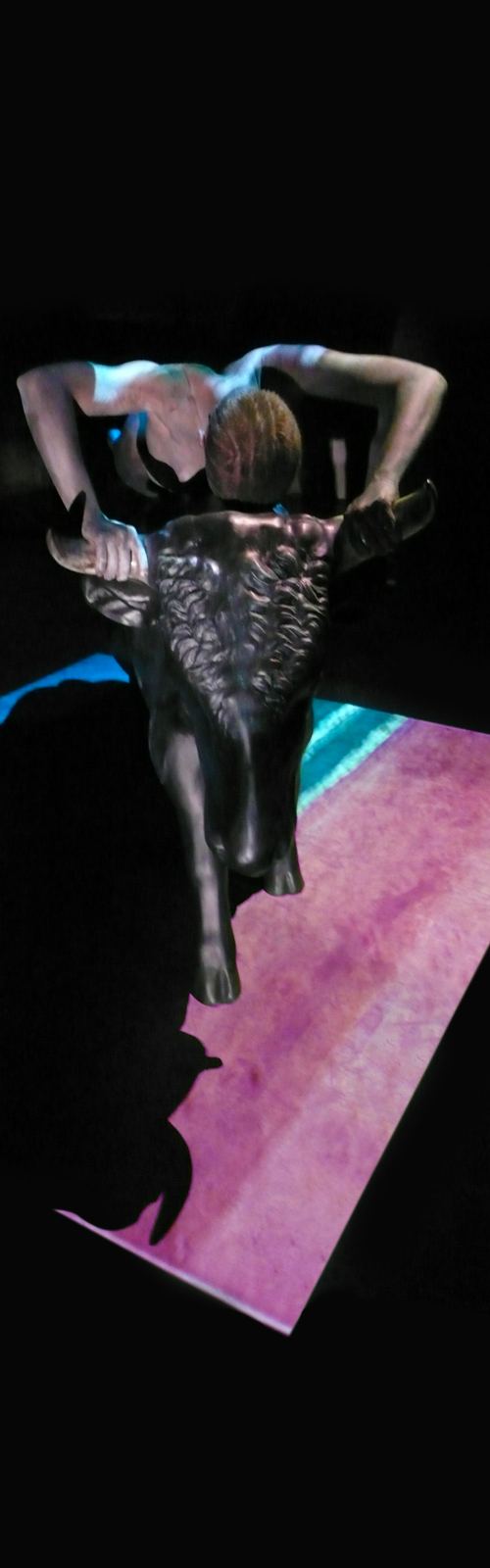 Sfondo nero, un corpo gracile cavalca la statua nera di un toro, ai suoi piedi un tappeto celeste e rosa carne.