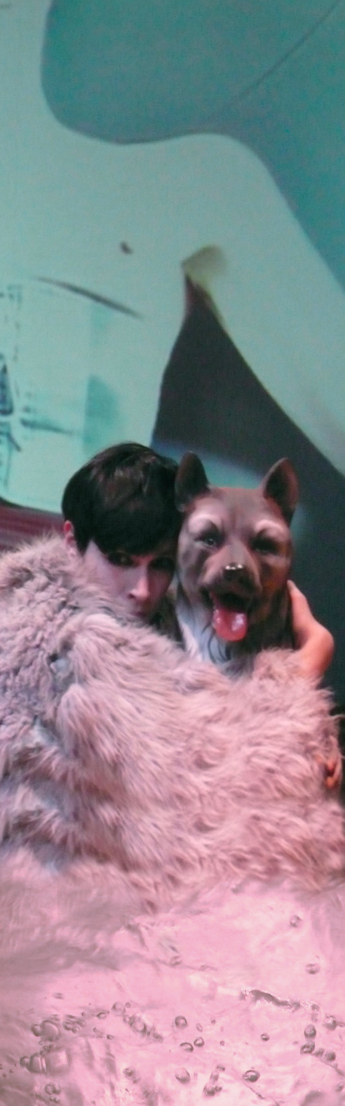 In the foreground a young woman wears a fur coat and hugs a statue of a dog, behind the video projection of the same young woman forced into a bodice that squeezes her chest so tightly until her breasts come out