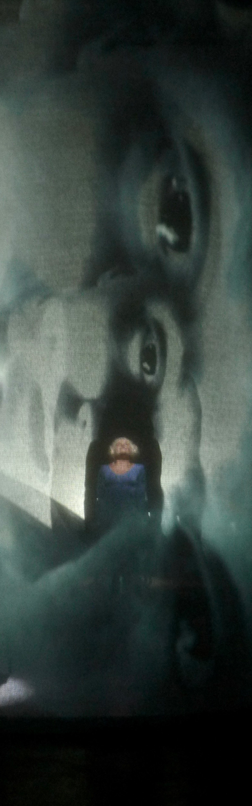 Sfondo nero, una proiezione fantasmatica di occhi e volti si sviluppa in verticale, al centro è riconoscibile una figura umana vestita di blu che guarda verso l'alto.